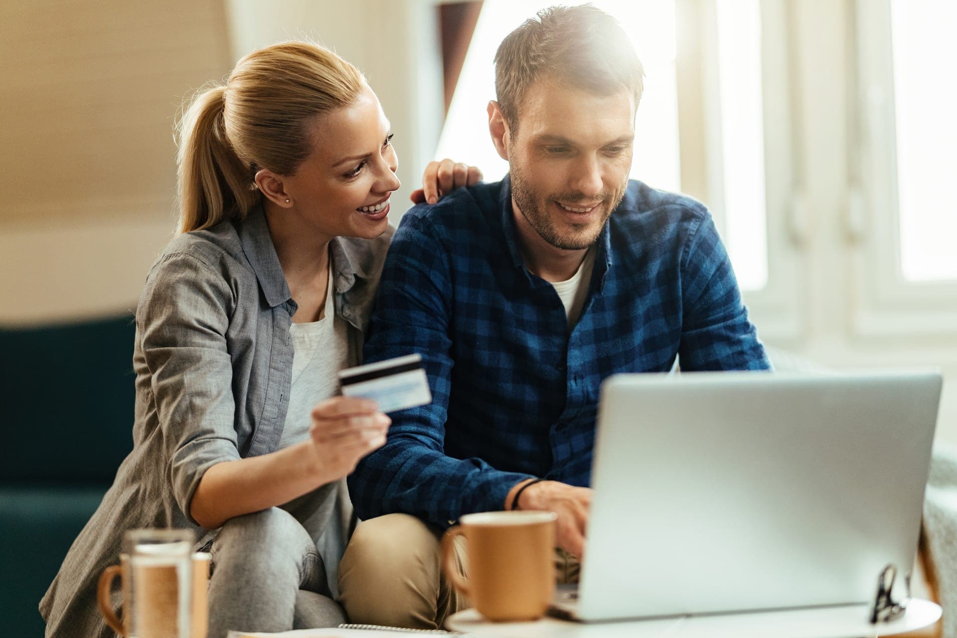 Een stel kijkt samen ontspannen naar een laptop terwijl ze met een bankpas in de hand een keuze vergelijken, met koffie op tafel